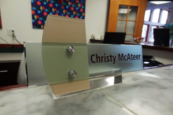 A nameplate on a marble counter reads "Christy McAteer." The background shows an office setting with a colorful painting, a wooden door with glass panes, and office chairs. Natural light is coming through windows.