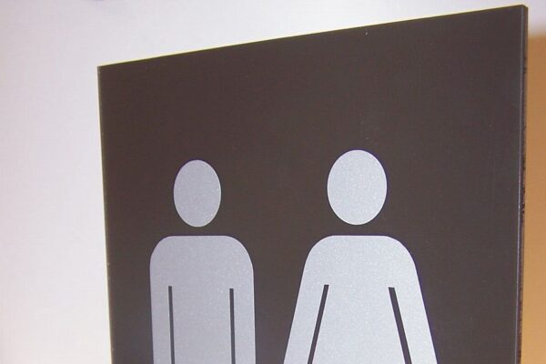 A rectangular bathroom sign mounted on a wall bracket with symbols indicating male and female restrooms. The sign features two simple white stick figures, one male and one female, on a dark background.