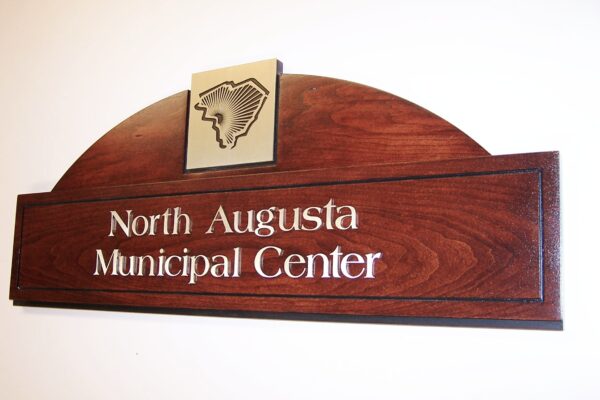 A wooden sign with an arched top displays the text "North Augusta Municipal Center" in white lettering. The sign also features an engraved outline of the state of South Carolina near the top.