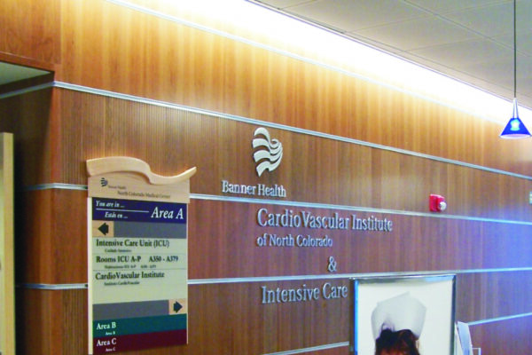 A medical facility hallway with wooden panel walls and a sign that reads "Cardio Vascular Institute of North Colorado & Intensive Care." A signboard lists different areas, and a large poster of a child dressed as a nurse is displayed near a desk.