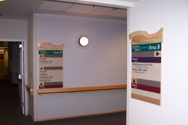 An interior hallway with two directory signs on the walls, labelled "Area B." Each sign lists multiple room numbers and departments in different colors. The walls are white and there is a circular light fixture in the middle of the wall.