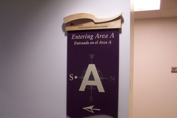 A sign on a wall reads "Entering Area A / Entras a el Area A" with a large letter "A" in the center and compass directions around it (N, E, S, W). An arrow pointing left is at the bottom of the sign. The top portion includes an arched decorative element.
