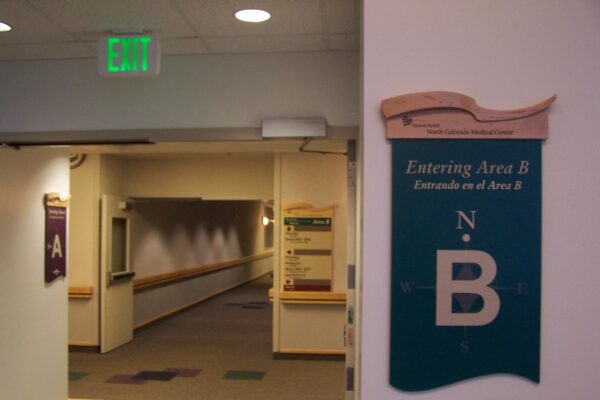 A hallway in a building with a sign indicating "Entering Area B" in English and Spanish. The sign features a compass symbol pointing north. Another sign for Area A is visible in the background. The hallway is well-lit with overhead lighting and an exit sign.
