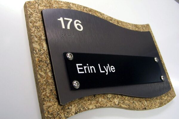 A wall-mounted, curved-edge nameplate with the number 176 at the top and 'Erin Lyle' written below on a black plaque. The border of the plaque has a textured, cork-like appearance.