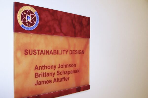 Plaque on a wall reading, "Sustainability Design" with names listed below: "Anthony Johnson, Brittany Schapanski, James Altaffer." The plaque has a red, marble-like background. The upper left corner features a logo with an atom design.