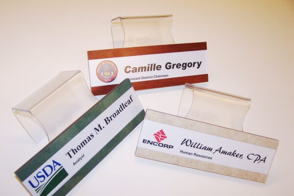 The image shows three desk nameplates. One is labeled "Camille Gregory" and has a logo on the left. Another reads "Thomas M. Broadleaf" with a USDA logo. The third nameplate is for "William Awaker, CPA" with an ENCORP logo. Each nameplate is on a clear stand.
