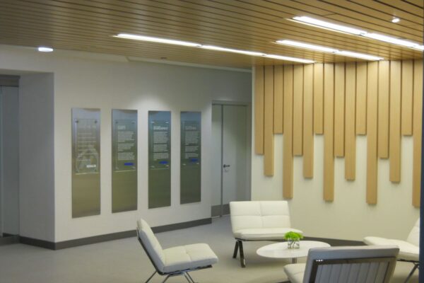 A modern office lobby with wooden paneling on the wall and a wooden ceiling. The lobby is furnished with white minimalist chairs and a small round white coffee table with a green decorative piece on it. Four framed pieces of art are displayed on the wall.