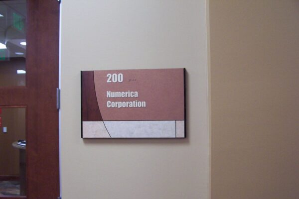 A sign on a beige wall reads "200 Numerica Corporation." The sign is rectangular with a maroon and white color scheme. A door with a glass panel is partially visible to the left of the sign.
