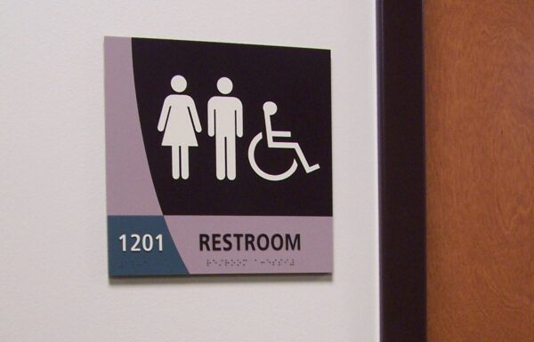 A restroom sign on a wall indicates a unisex and accessible restroom. The sign shows icons for a woman, a man, and a person in a wheelchair. The number 1201 is displayed above the word "Restroom.