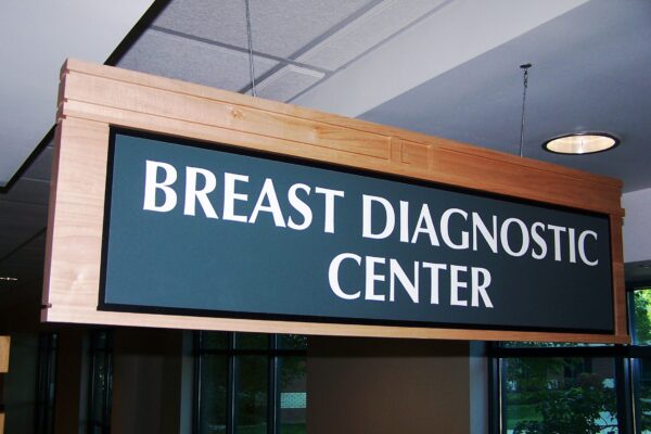 A rectangular wooden sign with a dark blue background and white text hangs from a ceiling, reading "BREAST DIAGNOSTIC CENTER." The sign is located indoors, with windows and fluorescent lights visible in the background.