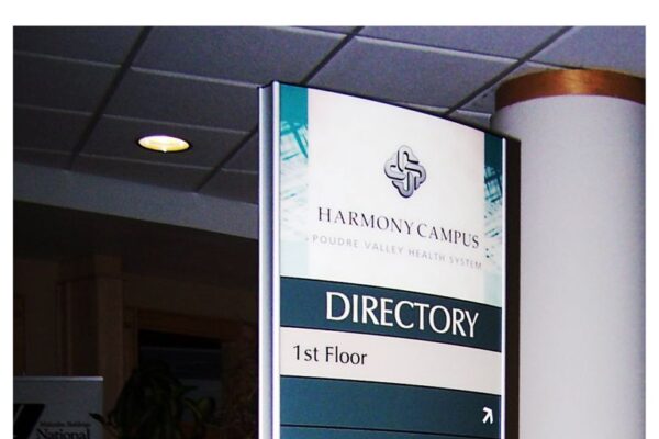 A standing directory sign in a medical facility lists locations for various departments: Imaging Center, Urgent Care Center, Breast Diagnostic Center, Harmony Café, Medical Offices, Elevator, and Harmony Surgery Center, with arrows indicating directions.