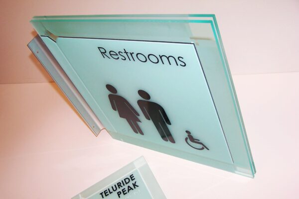A close-up image of a frosted glass sign for restrooms, depicting symbols for male, female, and wheelchair accessibility. The sign is mounted on a wall, with another smaller sign below it that reads "Telluride Peak." The background is white.