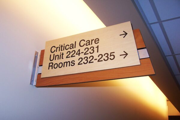 A hallway sign indicating directions to "Critical Care Unit 224-231" and "Rooms 232-235" with arrows pointing right. The sign is mounted on a wooden bracket on a lightly colored wall, and the ceiling features recessed lighting and ceiling tiles.
