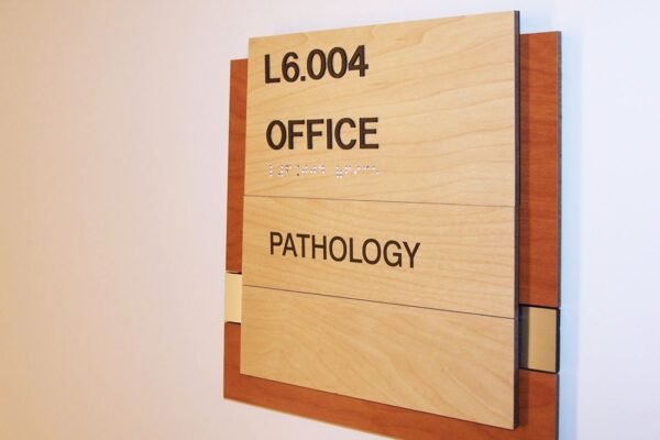 A wall-mounted office sign with a wooden frame and beige panels. The top panel reads "L6.004." The middle panel reads "OFFICE," and the bottom panel reads "PATHOLOGY.