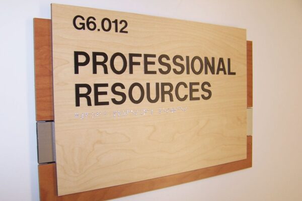 A wall-mounted wooden sign with the text "G6.012 PROFESSIONAL RESOURCES" in black letters. The sign has a light wood finish with two darker wood slats at the top and bottom edges.