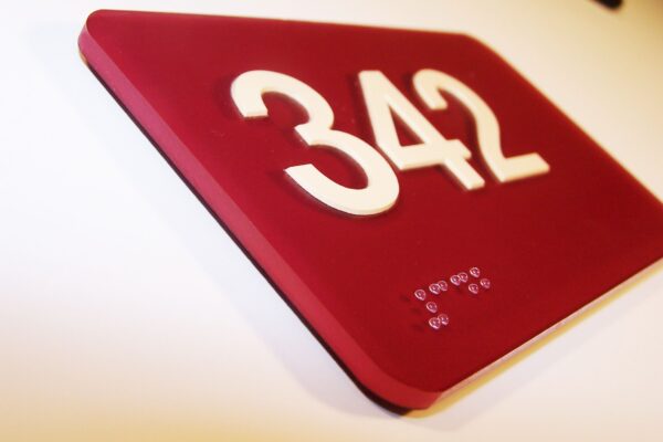 A red rectangular sign with the white number "342" prominently displayed. Below the number is a series of braille dots. The sign is mounted against a light-colored wall, slightly tilted upward.