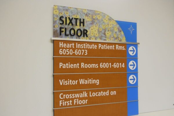 A wall-mounted sign indicating directions on the sixth floor of a building. It lists Heart Institute Patient Rooms 6050-6073, Patient Rooms 6001-6014, Visitor Waiting, and mentions a crosswalk located on the first floor, with arrows pointing right.