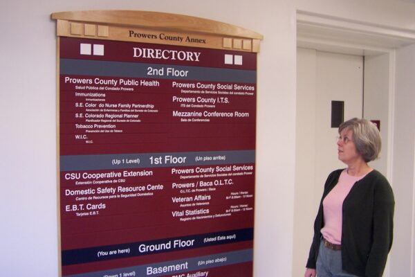 A woman with short gray hair and glasses stands looking at a large directory sign inside a building. The directory lists various departments and offices located on different floors, including a public health office, social services, and safety resource center.