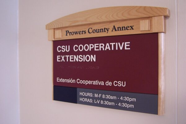 A sign on a wall reads "Prowers County Annex" at the top. Below, it says "CSU Cooperative Extension" and "Extensión Cooperativa de CSU." The hours of operation are listed as Monday to Friday, 8:30 am - 4:30 pm in both English and Spanish.