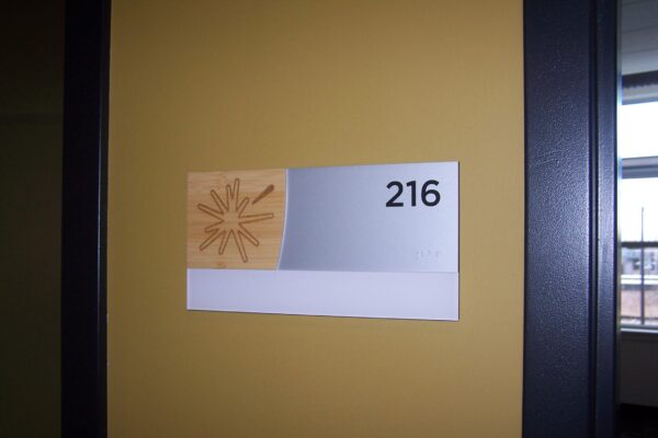 A room number sign on a yellow wall displays the number 216 in black text on a silver background. Beside the number is a design of abstract lines on a light brown background.