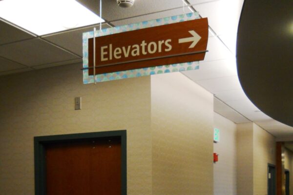 A hallway with a brown door on the left side and a large sign above pointing right labeled "Elevators." The corridor features gray floor tiles and light-colored walls. Several other doors are visible further down the hallway.