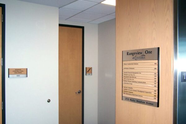 A hallway with off-white walls and a light wooden door. On the wall, there is a large directory sign listing various rooms and their locations. A smaller sign is mounted on the adjacent wall to the door, displaying additional information. The lighting is bright and even.