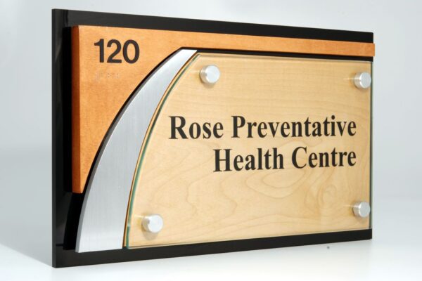 A wall sign with the number "120" in the upper left corner. The sign reads "Rose Preventative Health Centre" written on a light wood background, bordered by a black frame with a modern metal accent on the left-hand side.