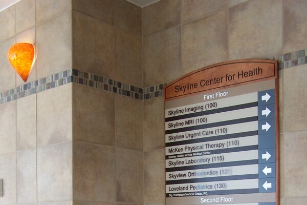 A directory sign mounted on a beige, tiled wall in a building with directions to various departments. A green plant in a pot sits at the base of the wall. An orange sconce light is visible above the sign. The area appears clean and well-maintained.