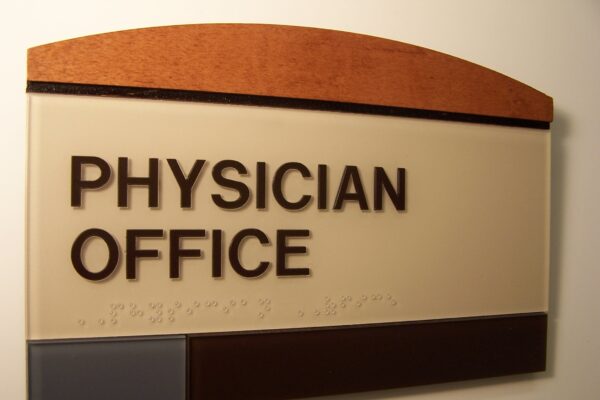 A wall-mounted sign with a wooden arch at the top reads "PHYSICIAN OFFICE" in large, bold letters. The sign consists of beige, brown, and gray panels with Braille text beneath the main lettering.
