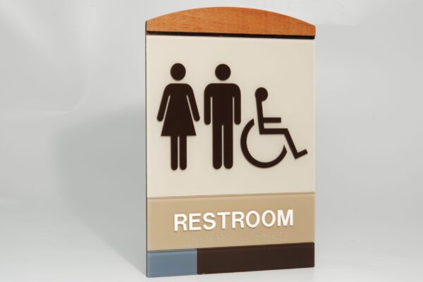 Sign indicating a restroom for multiple users. The sign has pictograms of a woman, a man, and a person in a wheelchair, above the text "RESTROOM". The top of the sign is curved and colored in brown and beige tones.