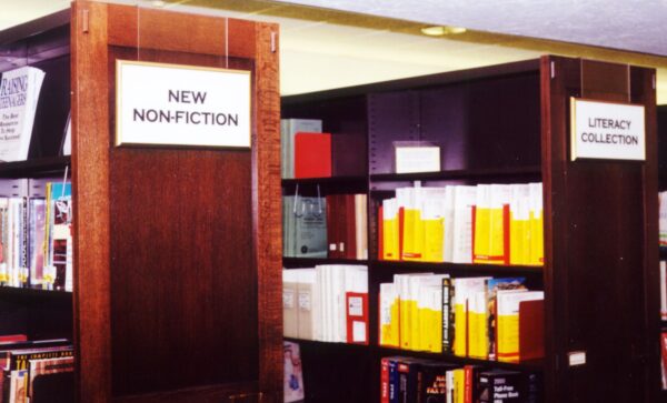 Two wooden bookshelves in a library. The left shelf, labeled "New Non-Fiction," is filled with various books. The right shelf, labeled "Literacy Collection," contains books with yellow spines and other materials.