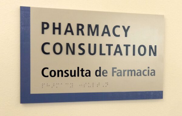 A sign on a wall says "PHARMACY CONSULTATION" in large bold letters, with "Consulta de Farmacia" in smaller text below. There is also Braille text at the bottom of the sign on a blue strip.