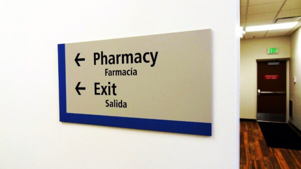 A directional sign in a hallway with arrows pointing left for "Pharmacy" (Farmacia) and "Exit" (Salida). The wall is white, and the hallway floor is wooden. There is a green exit sign above a brown door in the background.