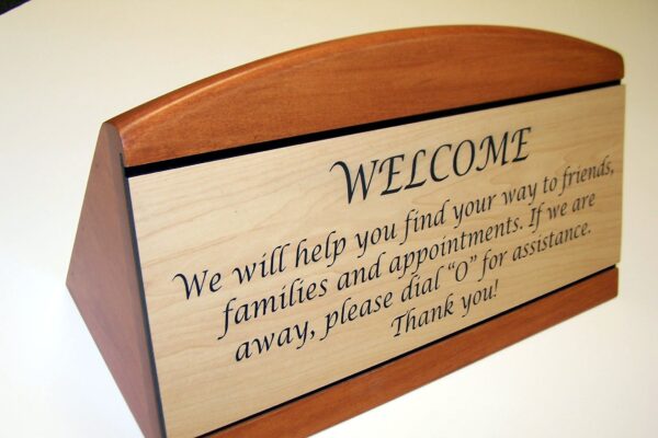A wooden desk sign with a light-colored front panel engraved with a welcome message in elegant script. The message reads, "WELCOME. We will help you find your way to friends, families and appointments. If we are away, please dial '0' for assistance. Thank you!.