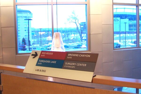 A directional sign inside a building points to various locations: to the right for Browns Canyon and Surgery Center/Orthopedic, and to the left for Red Rocks and Turquoise Lake. Large windows in the background reveal a snowy, urban landscape.