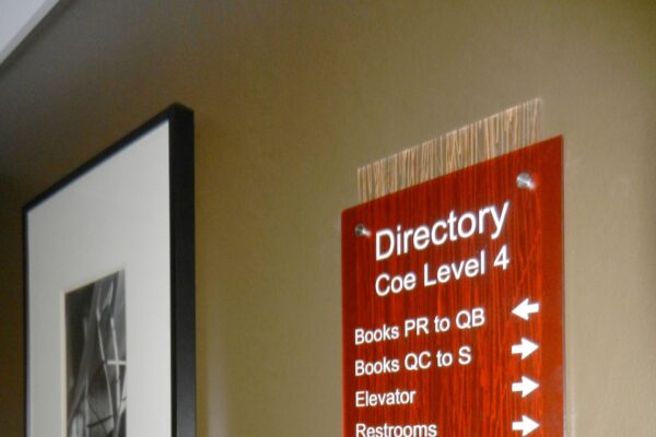 A wall-mounted directory sign on Coe Level 4 with arrows pointing to "Books PR to QB," "Books QC to S," "Elevator," "Restrooms," and "Study Room 463." Next to the sign is a framed black-and-white photograph.
