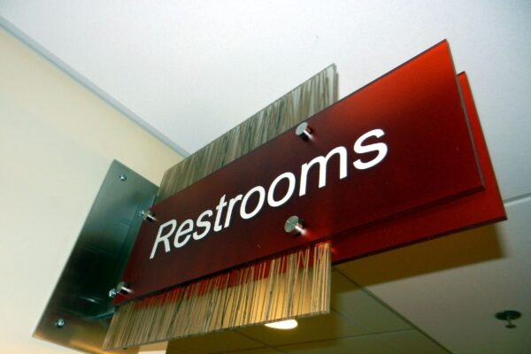 A directional sign for restrooms is mounted on a wall. The sign is red with white text that reads "Restrooms." The sign also includes a decorative element with vertical lines on the left side. It is lit from above by a ceiling light.