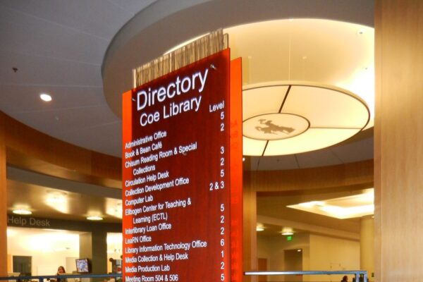 A vertical red directory sign with white text stands on a metal base in a spacious, modern building with multiple floors. The sign lists the locations of various offices and facilities within the Coe Library and features a wooden accent at the bottom.
