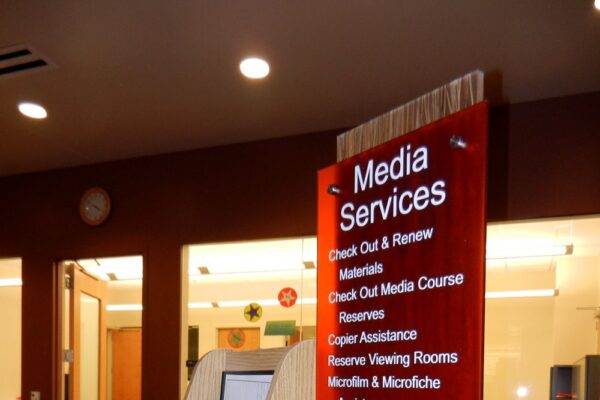 A library media services desk with a red sign listing available services including course reserves, media check-out and renewals, and group assistance. Brochures and information are displayed in holders on the counter. The background shows additional office space.