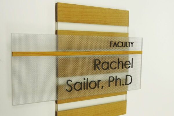 A wall-mounted sign with wood and frosted glass panels reads "FACULTY" at the top and "Rachel Sailor, Ph.D" below it in black text. The sign is placed on a white wall.
