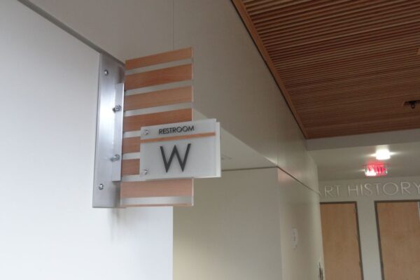 A restroom sign hangs on a wall, featuring the letter "W" to indicate the women's restroom. The sign is attached to a metal bracket and is positioned against a neutral-colored wall and ceiling with wooden panels. A lit exit sign and two closed doors are visible in the background.