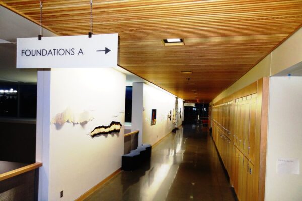 A brightly lit hallway with wooden ceiling and lockers on the right. The floor is polished concrete. Art pieces hang on the left wall, and a sign overhead reads "Foundations A" with an arrow pointing to the right.