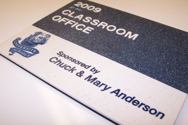 A sign with a silver and white background reads "2009 Classroom Office" and "Sponsored by Chuck & Mary Anderson." It features an emblem of an anchor and a banner with the words "Maritime Academy." The sign is placed against a white surface.