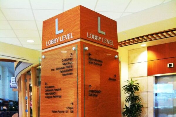A wooden directory pillar stands in a lobby area, featuring a large "L" at the top for "Lobby Level." The directory lists various departments and offices in black text on clear panels. The surroundings include a potted plant, elevator doors, and a glossy tiled floor.