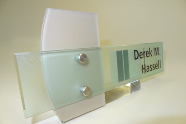 A translucent, modern nameplate with frosted glass and metallic accents reads "Derek M. Hassell." The nameplate features a contemporary design with a combination of clear and frosted glass segments, secured by two metal bolts.
