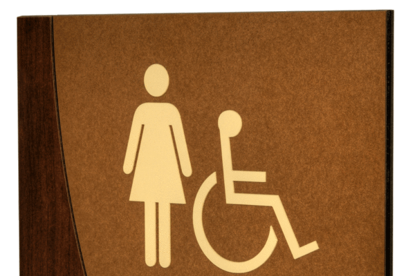 A restroom sign indicating facilities for women and individuals with disabilities. The sign features a female figure and a wheelchair symbol on a brown and beige background with the word "WOMEN" in bold text at the bottom.