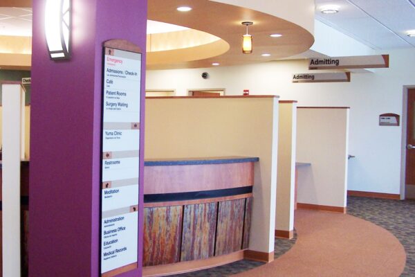 A modern lobby area with a purple pillar on the left that features a directory sign, a curved wooden reception desk in the center, and various office rooms around the perimeter. The flooring is a mix of carpet and tile, and overhead lighting is contemporary.