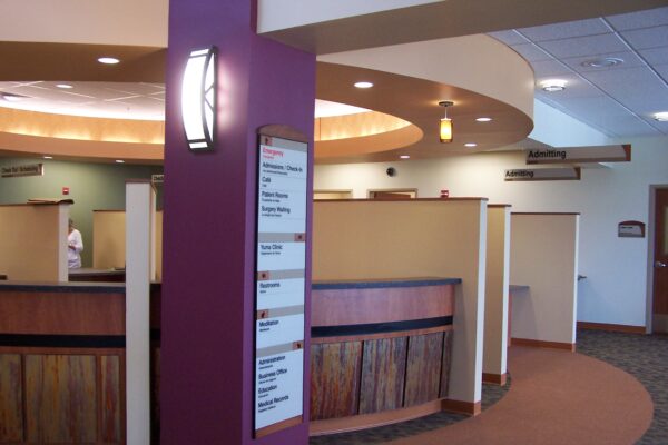 A modern medical reception area featuring a curved wooden desk with dividers, a purple column with a light fixture and a sign listing departments. Neutral-toned walls and carpeting provide a calming atmosphere. Signs point to various sections of the facility.