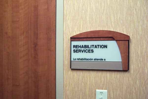 A sign on a beige wall reads "REHABILITATION SERVICES" in both English and Spanish, with braille writing underneath. The sign has a transparent top section and a wooden frame.