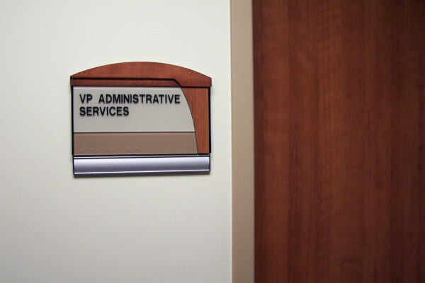 A wall-mounted sign next to a wooden door reads "VP Administrative Services." The sign has a brown and beige color scheme with a silver bottom section. The door is partially visible with a metal handle.